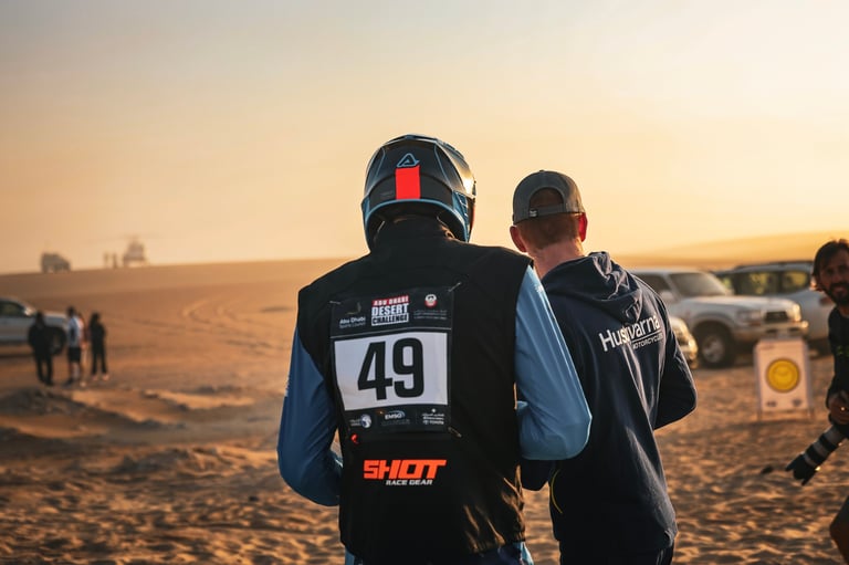 Motorcyclist wearing race number 49 and helmet standing in desert at sunset with spectators in background