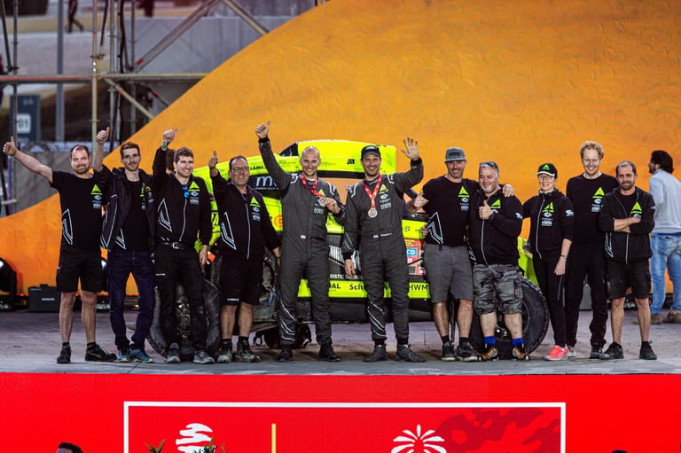 Team members in black and neon yellow vests celebrating with raised arms on a red podium in front of a large orange dome