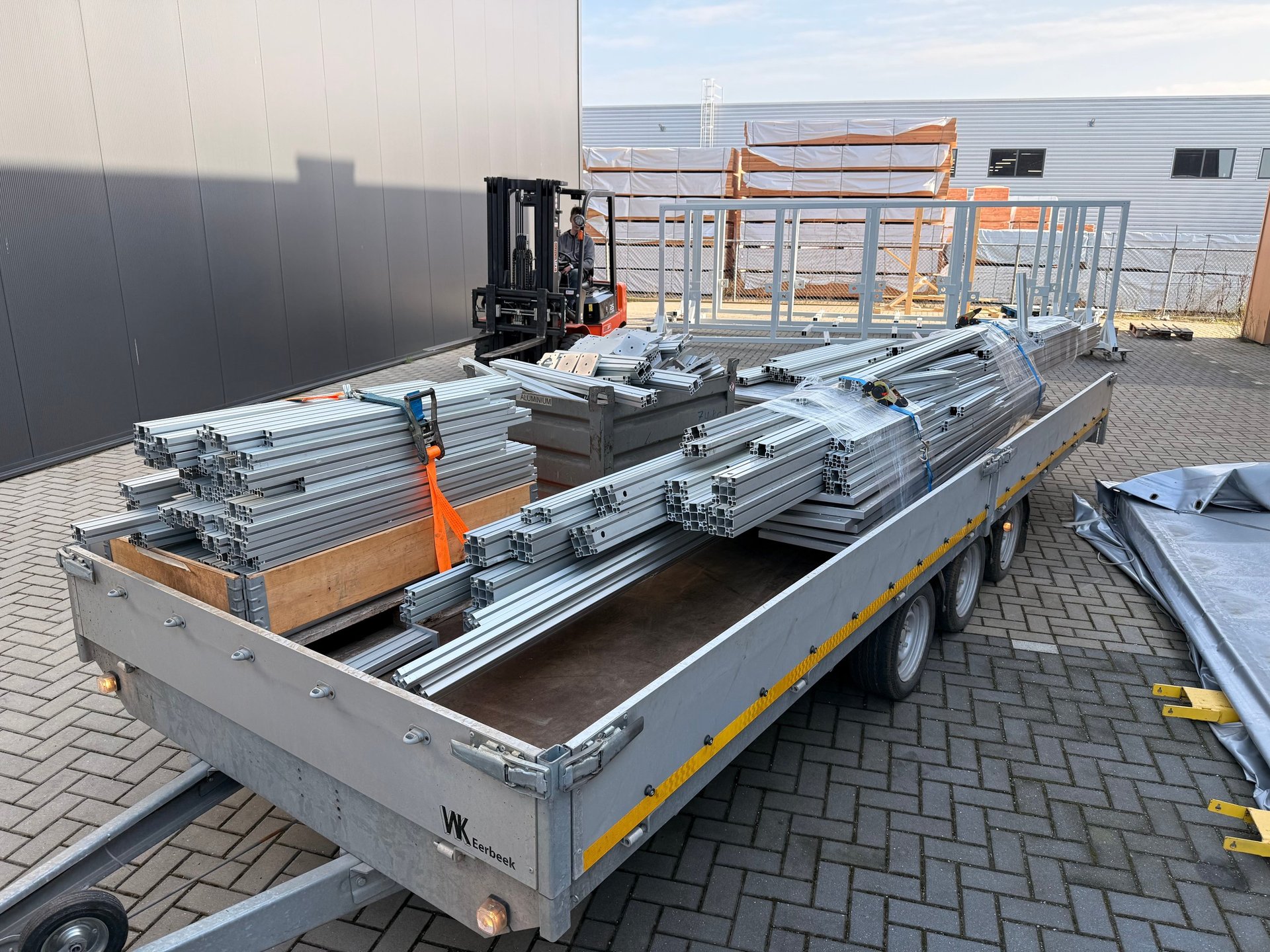 Flatbed trailer loaded with stacked metal materials and equipment, parked in an industrial yard with buildings and forklifts visible.