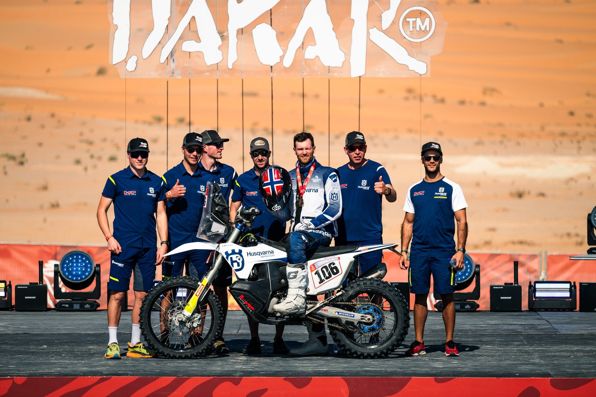 Dakar rally team posing with white and black motorcycle number 108 on stage with orange desert backdrop